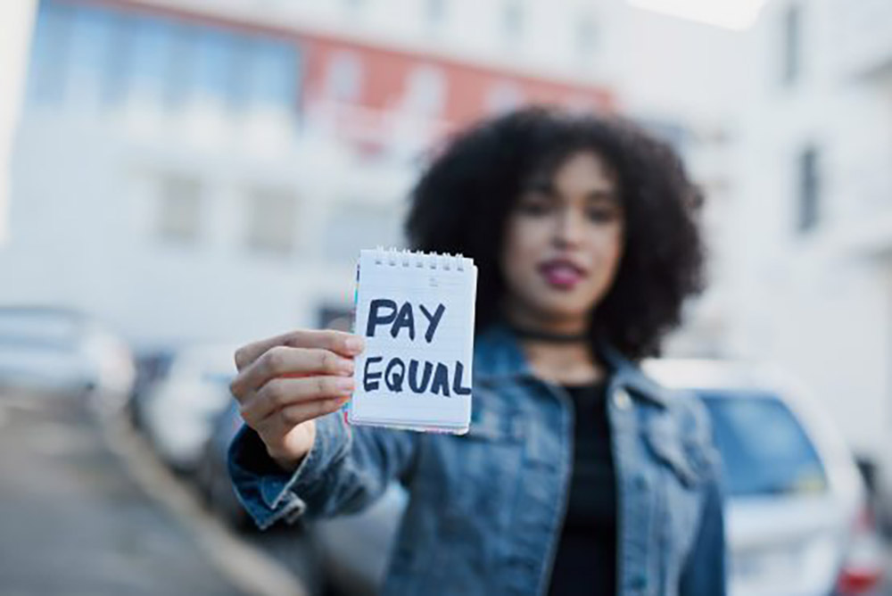 Someone holds a piece of paper reading 'Pay Equal'