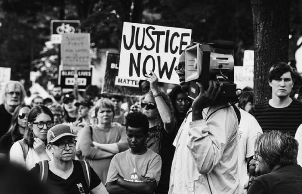 black and white image of a protest, with a sign reading 'Justice Now'